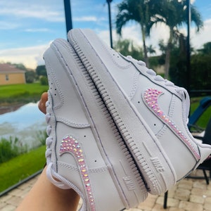 Puede incluir: Zapatillas blancas de cuero con un dise&ntilde;o de swoosh de cristal rosa e iridiscente.