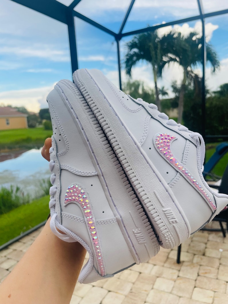 Puede incluir: Zapatillas blancas de cuero con un dise&ntilde;o de swoosh de cristal rosa e iridiscente.