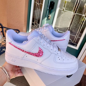 Puede incluir: Zapatillas bajas de cuero blanco con un swoosh Nike de pedrer&iacute;a rosa y detalles de pedrer&iacute;a rosa en los cordones. Las zapatillas est&aacute;n sobre una caja blanca con la palabra "AIR" impresa en el lateral.