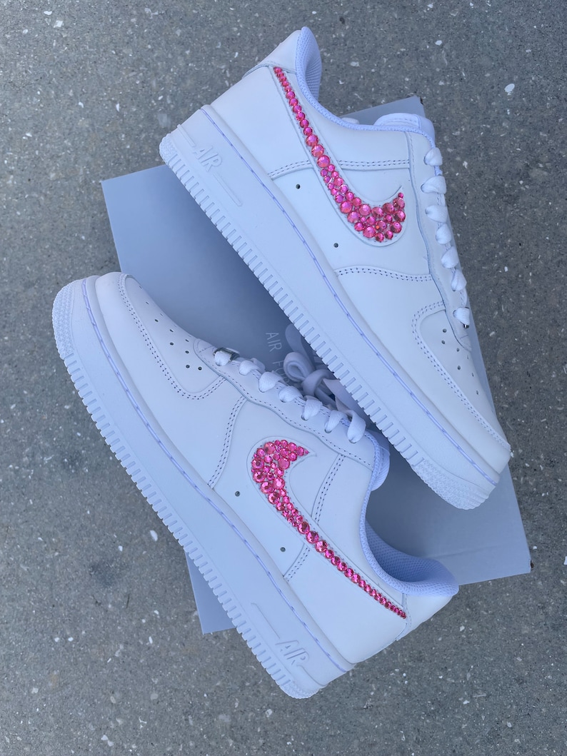 Puede incluir: Zapatillas blancas de cuero con un swoosh Nike de diamantes de imitaci&oacute;n rosa. Las zapatillas est&aacute;n sobre una caja blanca con la palabra "AIR" impresa en el lateral.