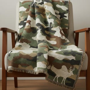 Puede incluir: Una manta con estampado de camuflaje sobre un sill&oacute;n de madera. La manta presenta un patr&oacute;n repetido de siluetas de ciervos en tonos verdes, marrones y beige. La manta tiene un borde con flecos.