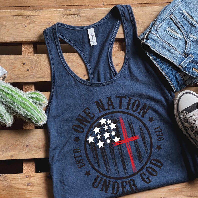 May include: A navy blue tank top with a white and red graphic design. The graphic features a circle with the text "One Nation Under God Est. 1776" and a stylized American flag with a red cross.