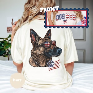 May include: Ivory t-shirt with a graphic of a German Shepherd wearing sunglasses and a bandana with a paw print and American flag. The text "Dog Mom" is visible on the back of the shirt.