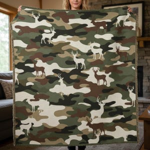 Puede incluir: Una manta con estampado de camuflaje con un patr&oacute;n repetido de siluetas de ciervos blancos y marrones sobre un fondo de formas verdes, marrones y crema. La manta tiene un borde con flecos y est&aacute; sujeta por una persona.