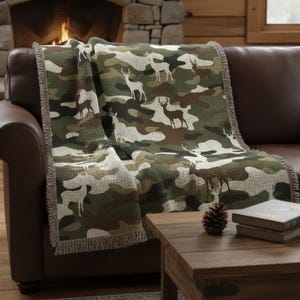 Puede incluir: Una manta con estampado de camuflaje con siluetas de ciervos, sobre un sof&aacute; de cuero marr&oacute;n. El patr&oacute;n de la manta incluye tonos verdes, marrones y blancos. Tiene un borde con flecos. Una mesa de centro de madera est&aacute; en primer plano.