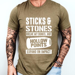 Può includere: Una maglietta verde con il testo "Sticks & Stones Break My Bones, But Hollow Points Expand On Impact" in lettere bianche. Il testo è stilizzato per assomigliare a un proiettile.