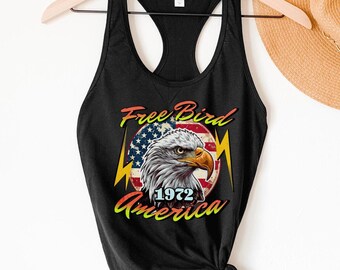 May include: Black tank top with a graphic design featuring an American bald eagle, the text "Free Bird," and the word "America" with the year "1972." The eagle is set against a backdrop of the American flag.