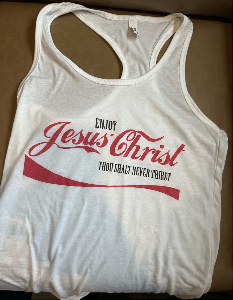 May include: White tank top with a red Coca-Cola-inspired graphic that says "Enjoy Jesus Christ Thou Shalt Never Thirst".