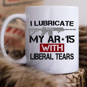May include: White ceramic coffee mug with a black and gray image of a rifle and the text "I lubricate my AR-15 with liberal tears".