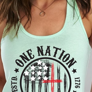 May include: A light green tank top with a black and red circle graphic. The graphic features a stylized American flag with a red cross and the text "ONE NATION UNDER GOD ESTD. 1776".