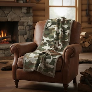Puede incluir: Un sill&oacute;n de cuero marr&oacute;n con una manta estampada con camuflaje que presenta siluetas de ciervos. La manta tiene un ribete de flecos y est&aacute; colocada sobre la silla. Un libro descansa sobre la manta. La escena est&aacute; ambientada en un interior r&uacute;stico, tipo caba&ntilde;a.