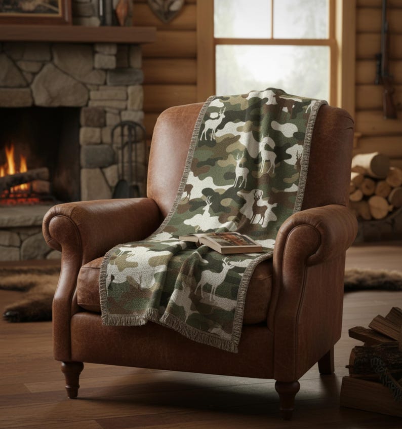 Puede incluir: Un sill&oacute;n de cuero marr&oacute;n con una manta estampada con camuflaje que presenta siluetas de ciervos. La manta tiene un ribete de flecos y est&aacute; colocada sobre la silla. Un libro descansa sobre la manta. La escena est&aacute; ambientada en un interior r&uacute;stico, tipo caba&ntilde;a.