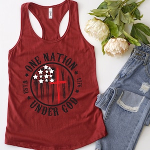 May include: A maroon tank top with a black and white graphic of an American flag with a red cross in the center. The graphic is surrounded by a circle with the text "ONE NATION UNDER GOD ESTD. 1776".