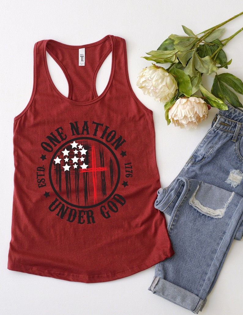 May include: A maroon tank top with a black and white graphic of an American flag with a red cross in the center. The graphic is surrounded by a circle with the text "ONE NATION UNDER GOD ESTD. 1776".