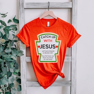 May include: Orange t-shirt with a ketchup-style graphic that says "Catch Up With Jesus." The graphic includes the text "Lettuce Praise & Relish Him" and "Cuz He loves me from my head to-ma-toes."