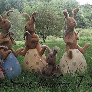 May include: A collection of brown rabbit figures, each with long ears, are positioned on decorative Easter eggs. Some rabbits hold paintbrushes. The eggs are painted in various colours and patterns, set in a grassy outdoor scene. The text "Sweet Meadows Farm" is at the bottom.