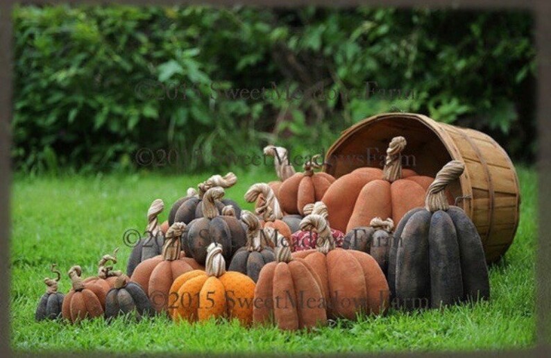 May include: A group of handmade fabric pumpkins in various shades of brown, orange, and black, spilling out of a wooden basket. The pumpkins are arranged on a bed of green grass.