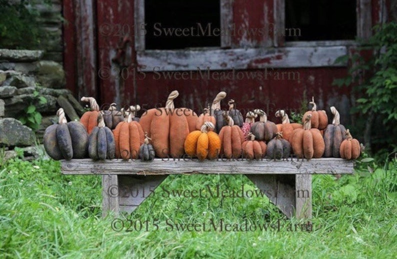 May include: A rustic wooden bench with a row of handmade fabric pumpkins in various sizes and colors. The pumpkins are a mix of orange, brown, and black, and some have stems. The bench is in front of a red barn with a wooden door.