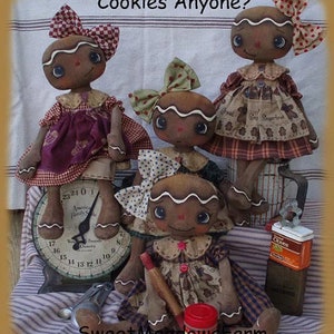 May include: Four gingerbread doll ornaments with brown eyes, red and white gingham dresses, and brown hair. The dolls are standing on a scale with a red and white striped tablecloth in the background. The text "Cookies Anyone?" is written above the dolls. The text "Sweet Meadows Farm" is written below the dolls.