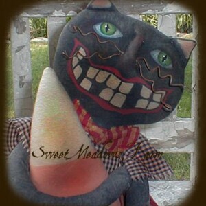 May include: A black cat with a wide, toothy grin and green eyes is wearing a plaid shirt and holding a large, white and orange candy corn. The cat is sitting on a wooden surface with a white picket fence in the background. The text "Sweet Meadows Farm" is written on the candy corn.