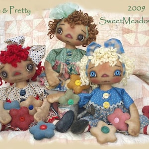 May include: Three handmade rag dolls with painted faces and yarn hair. The dolls are dressed in colorful floral print dresses and holding fabric flowers. The dolls are sitting on a white and pink patchwork quilt. The text "Prim & Pretty" and "Sweet Meadows Farm" is visible in the image.