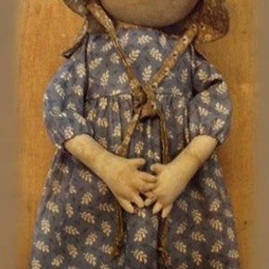 Puede incluir: Una muñeca de trapo hecha a mano con un vestido floral azul y blanco y un gorro marrón. La muñeca tiene un aspecto sencillo y rústico y está hecha de materiales naturales.