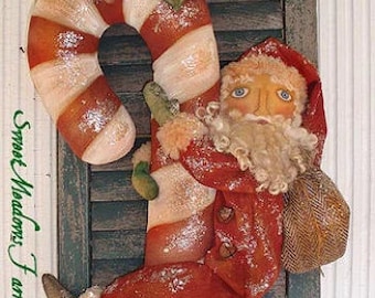 Puede incluir: Una decoración navideña hecha a mano con un muñeco de Papá Noel sujetando un gran bastón de caramelo con una rama de acebo en la parte superior. El bastón de caramelo es rojo y blanco con un acabado brillante. El muñeco de Papá Noel lleva un traje rojo y blanco con barba blanca y un sombrero marrón.