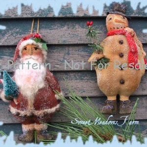 Op de afbeelding: Een handgemaakt vilt kerstman ornament met een rode muts, witte baard en bruine jas. Hij houdt een kleine groene boom en een snoepstok vast. Een sneeuwpop ornament met een rode sjaal en zwarte hoed is ook afgebeeld. De ornamenten worden getoond tegen een rustieke houten achtergrond.