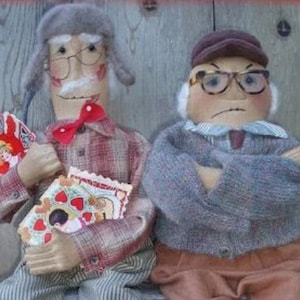 May include: Two handmade fabric dolls dressed as elderly men. One doll is wearing a gray hat, a red bow tie, and a plaid shirt. The other doll is wearing a brown hat, a gray sweater, and brown pants. Both dolls are holding Valentine's Day cards.