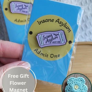 May include: Two blue rectangular cards with a yellow circular sticker that reads "Insane Asylum" and "Admit One". The cards are in clear plastic packaging. A small, crocheted blue flower is also visible.