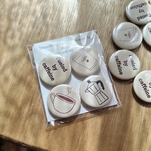 May include: A set of four circular pin-back buttons packaged in a clear plastic sleeve, featuring cream-colored backgrounds with dark brown line art of coffee cups, a moka pot, and the text "Fueled by caffeine" printed on the button surfaces.