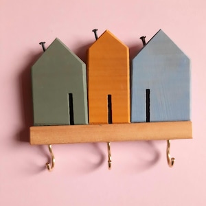 May include: A wooden key holder with three house-shaped cutouts in green, orange, and blue. Each house has a black door and a nail at the top. Three brass hooks are attached to a wooden base below the houses.