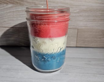 May include: A layered candle in a clear glass mason jar with a single wick. The wax is poured in three distinct horizontal stripes of red, white, and blue. The glass jar features the embossed word hello and small star shapes on the exterior surface.