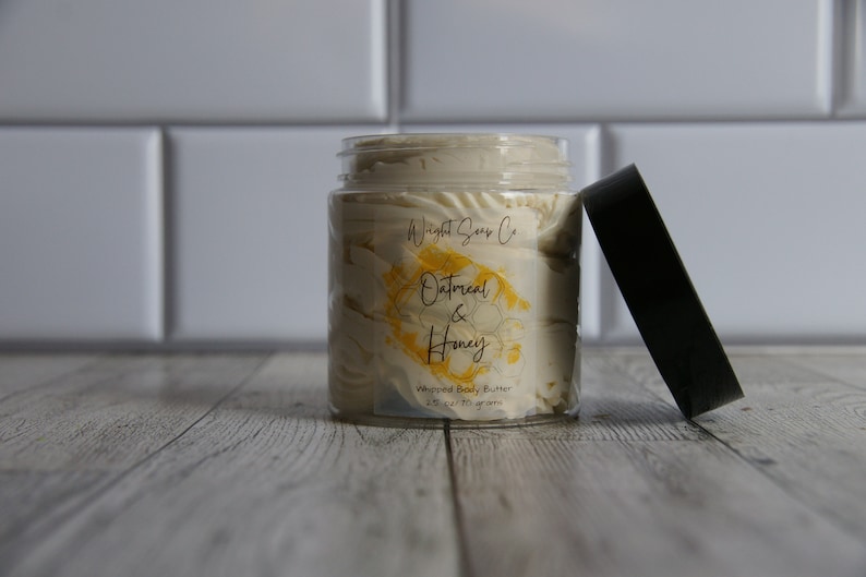 K&ouml;nnte beinhalten: Ein transparentes Glas mit wei&szlig;er, geschlagenem K&ouml;rperbutter mit einem gelben und wei&szlig;en Etikett, auf dem "Whipped Soap Co. Oatmeal & Honey Whipped Body Butter 2 oz / 57 grams" steht. Das Glas ist offen und der Deckel ist schwarz.
