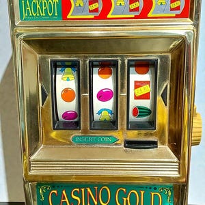 May include: A vintage gold-colored slot machine with the text "AUTOMATIC JACKPOT" and "CASINO GOLD". The reels display fruit and bell symbols. A red light sits atop the machine. The text "BELLS RING...LIGHT FLASHES...ON THESE COMBINATIONS!" is visible.