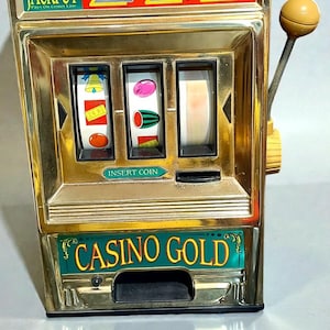 May include: Vintage gold-colored slot machine with the text "AUTOMATIC JACKPOT" and "CASINO GOLD". The machine has three reels with fruit symbols and a red light on top. It features a lever and a coin slot.