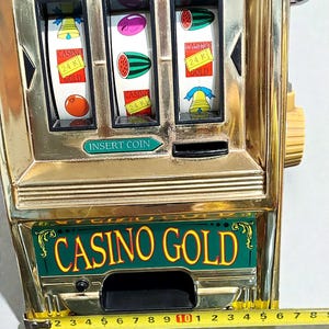 May include: A vintage gold-colored slot machine with three reels displaying fruit and bell symbols. The machine has the text "CASINO GOLD" and "24 K GOLD". A ruler shows measurements in inches.