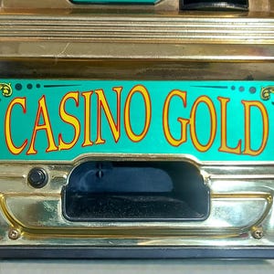 May include: Close-up of a vintage slot machine with a gold-colored frame. The machine features the text "CASINO GOLD" in yellow letters on a teal background. The reels display fruit symbols and the text "24K GOLD". The words "INSERT COIN" are also visible.