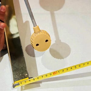 May include: A vintage, gold-toned metal object with a rounded top and a measuring tape showing inches. The object has a cylindrical stem and a circular, ridged base. The measuring tape shows approximately 4 inches.