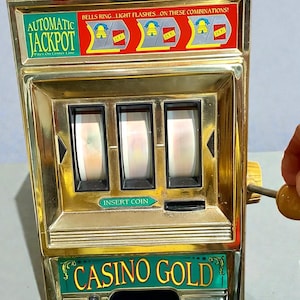 May include: A vintage toy slot machine with a gold-colored frame and a red light on top. The machine has three reels and the words "AUTOMATIC JACKPOT" and "CASINO GOLD" are visible. The text "INSERT COIN" is also displayed.