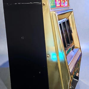 May include: A vintage slot machine with a black body and gold-colored trim. The machine has a red light on top and a display that reads "ULTIMATE JACKPOT". The machine has a classic design.