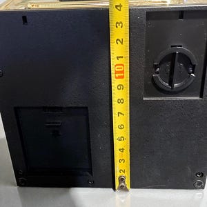 May include: A black and gold box-shaped object with a yellow measuring tape next to it. The box is approximately 4 inches tall. The measuring tape shows the height of the box. The box has a coin slot and a small door.