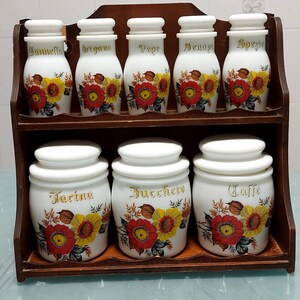 Può includere: Un porta spezie in legno a due ripiani con dieci barattoli in ceramica bianca con disegni floreali. I barattoli sono etichettati con lettere dorate: "Zannelli", "Origano", "Pepe", "Denoyo", "Spezie", "Farina", "Zucchero" e "Caffe".