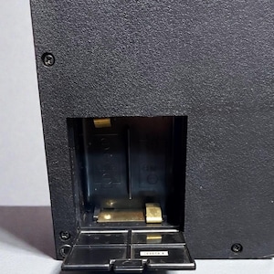 May include: Close-up of a black electronic device with a textured surface. The device has a battery compartment with an open door, revealing two slots. The device has a small, rectangular opening and is secured with screws.