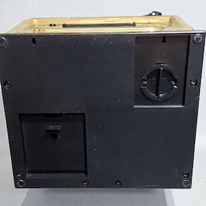 May include: A black rectangular box with a gold-colored top. The box has a battery compartment and a coin slot. The back of the box has a round opening with a twist-off cover. The box is likely a money bank or storage container.