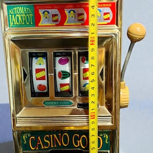 May include: A gold-colored toy slot machine with the words "AUTOMATIC JACKPOT" and "CASINO GOLD" on it. The machine has three reels with fruit symbols and a lever. The machine is approximately 7 inches tall.