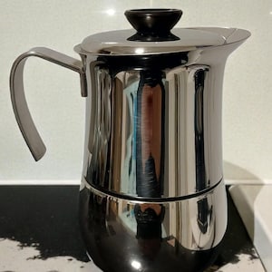 Puede incluir: Una taza de cerámica blanca con un asa negra y el texto "But First, Coffee" en negrita negra. La taza se encuentra sobre un fondo borroso, resaltando su diseño limpio.