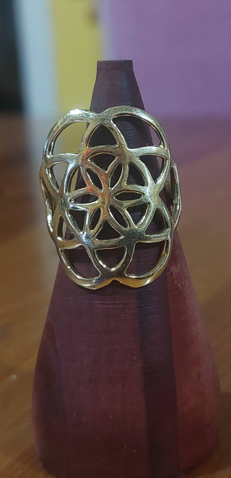 Puede incluir: Un anillo de color dorado con un dise&ntilde;o geom&eacute;trico que se asemeja a una flor. El anillo se muestra en un soporte de anillo de madera.