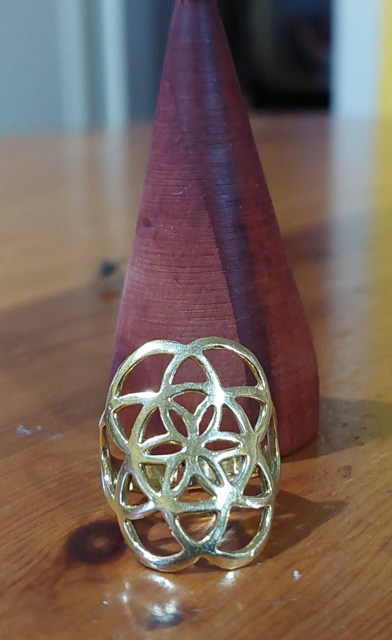 Puede incluir: Un anillo de color dorado con un dise&ntilde;o geom&eacute;trico que se asemeja a una flor. El anillo est&aacute; sobre una superficie de madera.