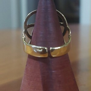 Puede incluir: Un anillo de tono dorado con un dise&ntilde;o geom&eacute;trico. El anillo se muestra en un soporte de anillo de madera.